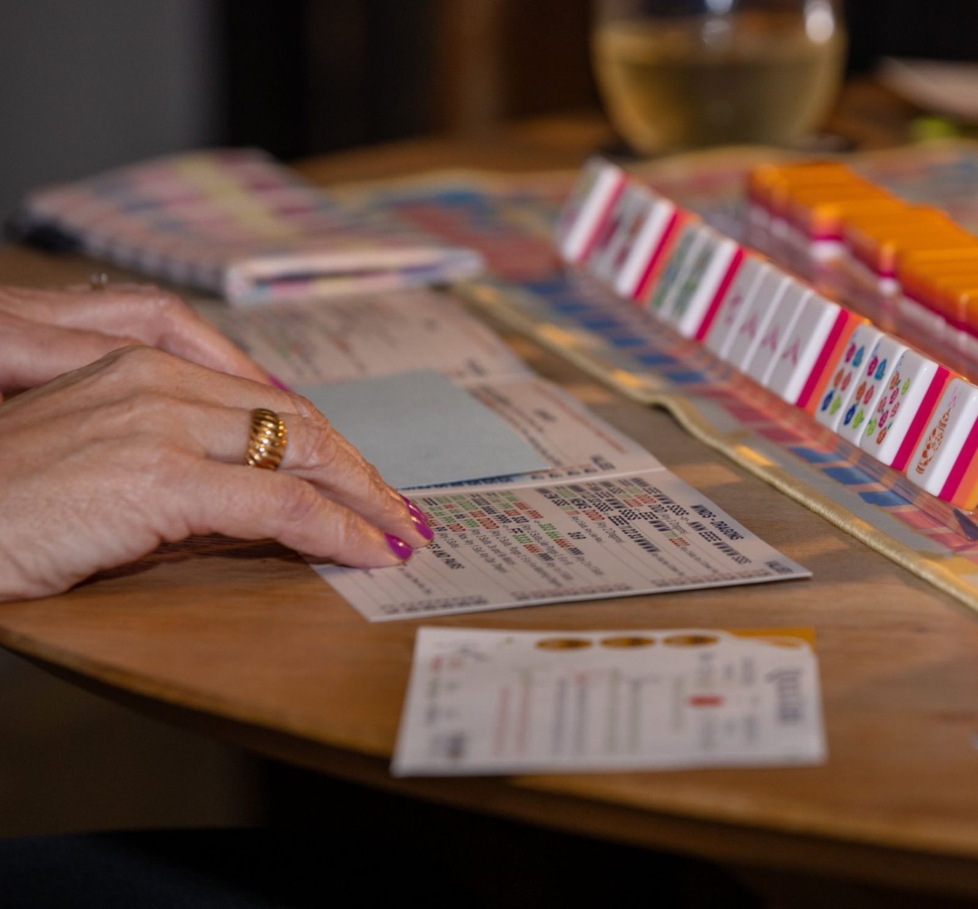 Hands resting on a mahjong card beside a rack of colorful pink and orange tiles and a glass of white wine