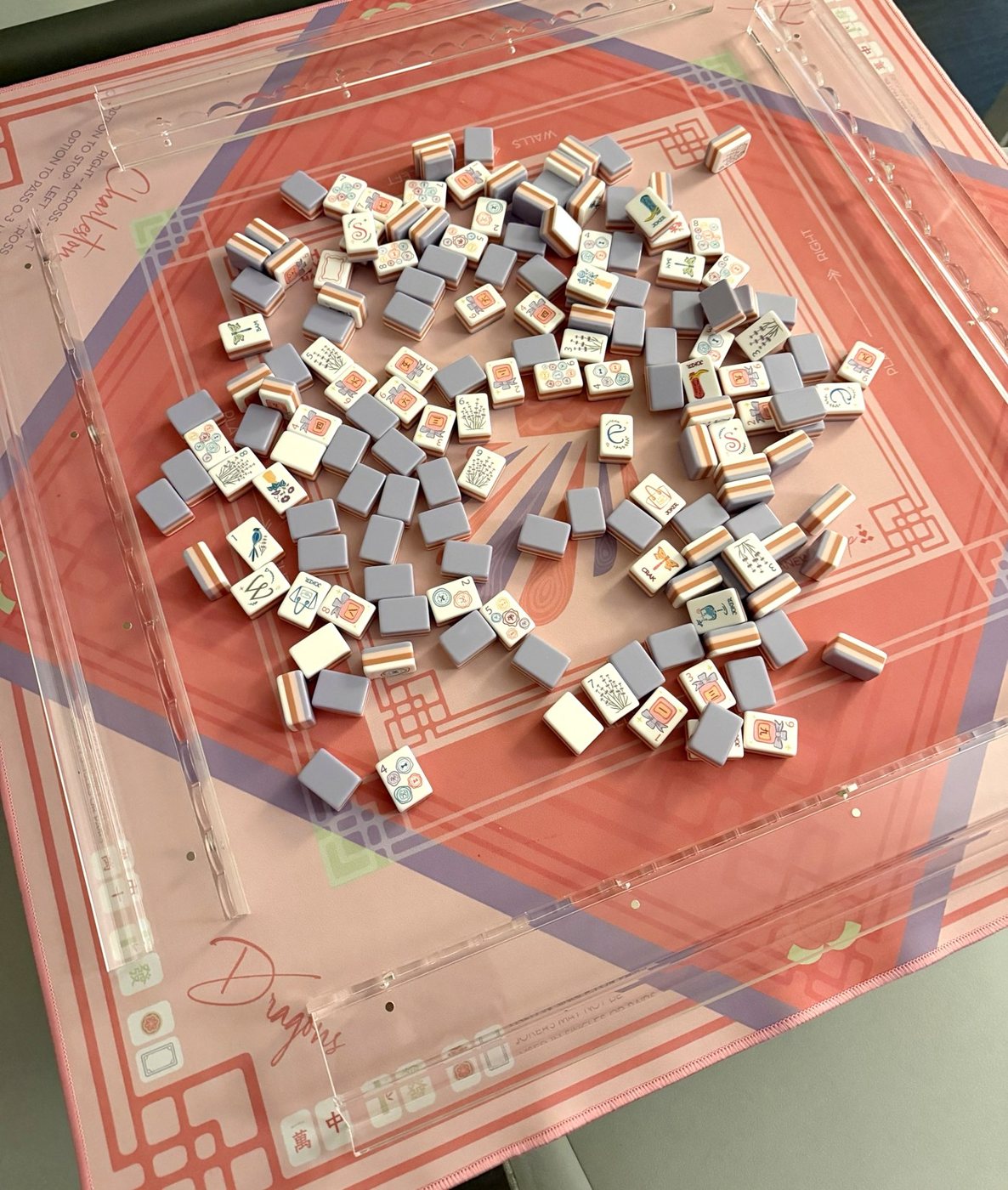A purple and white mahjong set spread out on a pink mahjong mat, framed by clear acrylic racks
