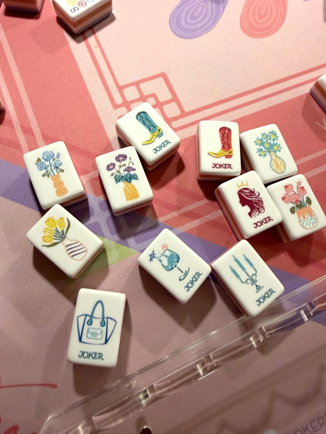 Mahjongolé joker tiles, including a cowboy boot, a wine glass, a handbag and a candelabra, with flower tiles on a pink mahjong mat