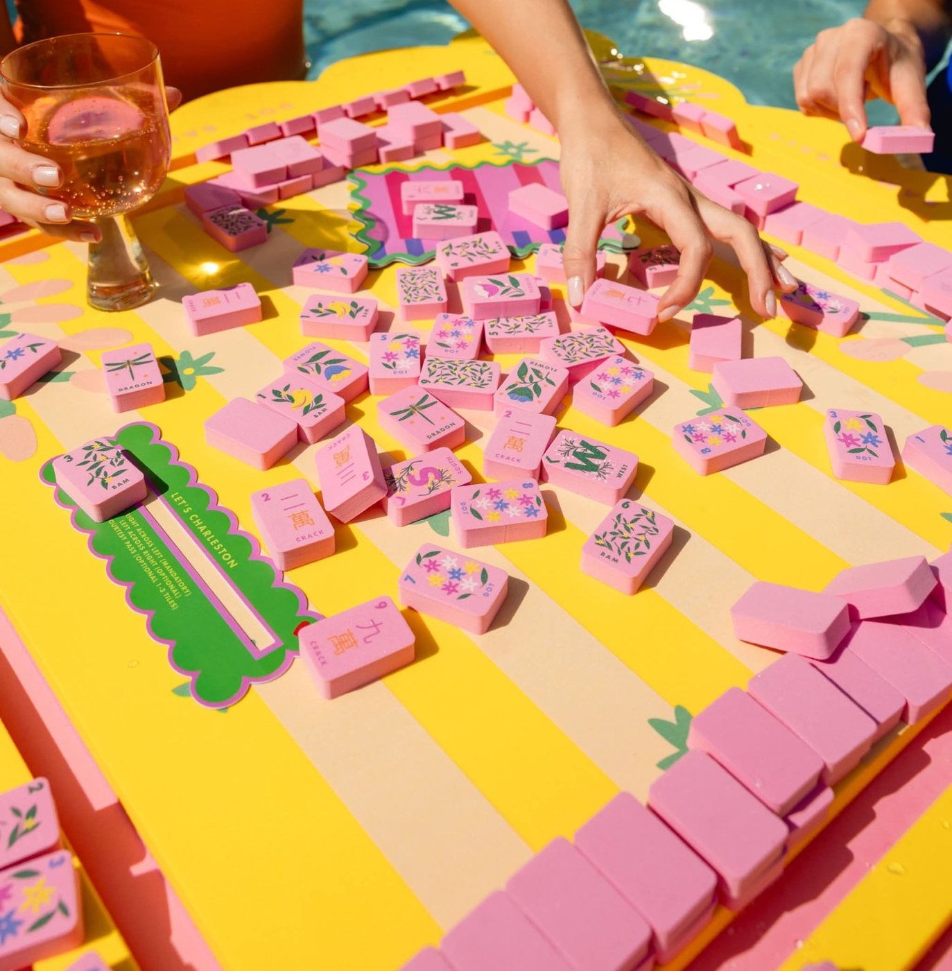 Friends playing with pink floral floating mahjong tiles on a yellow striped floating mat in a pool, one holding a glass of rosé