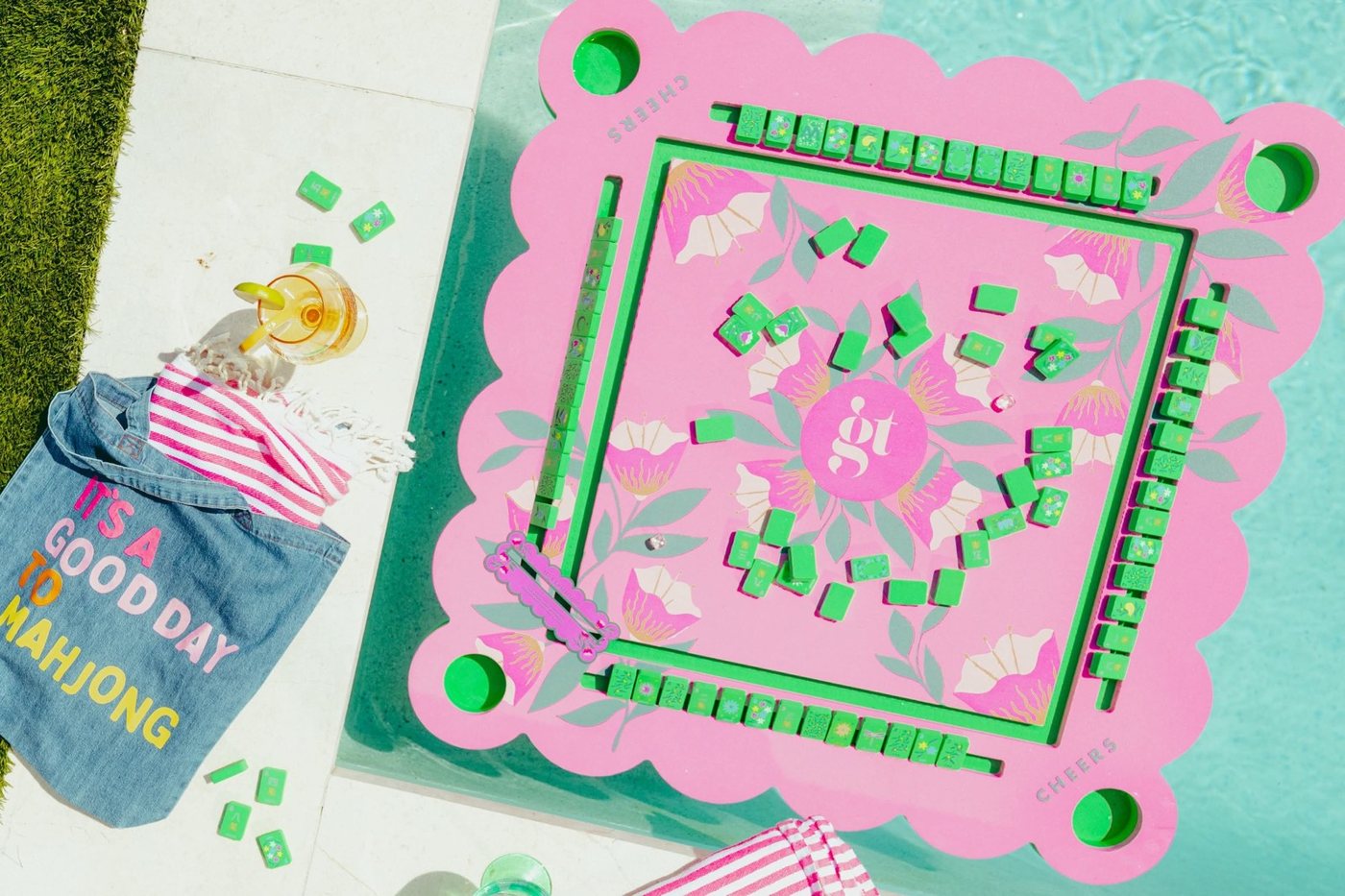 A pink scalloped floating mahjong mat with green tiles in a pool, beside a denim It's a Good Day to Mahjong tote and a striped towel