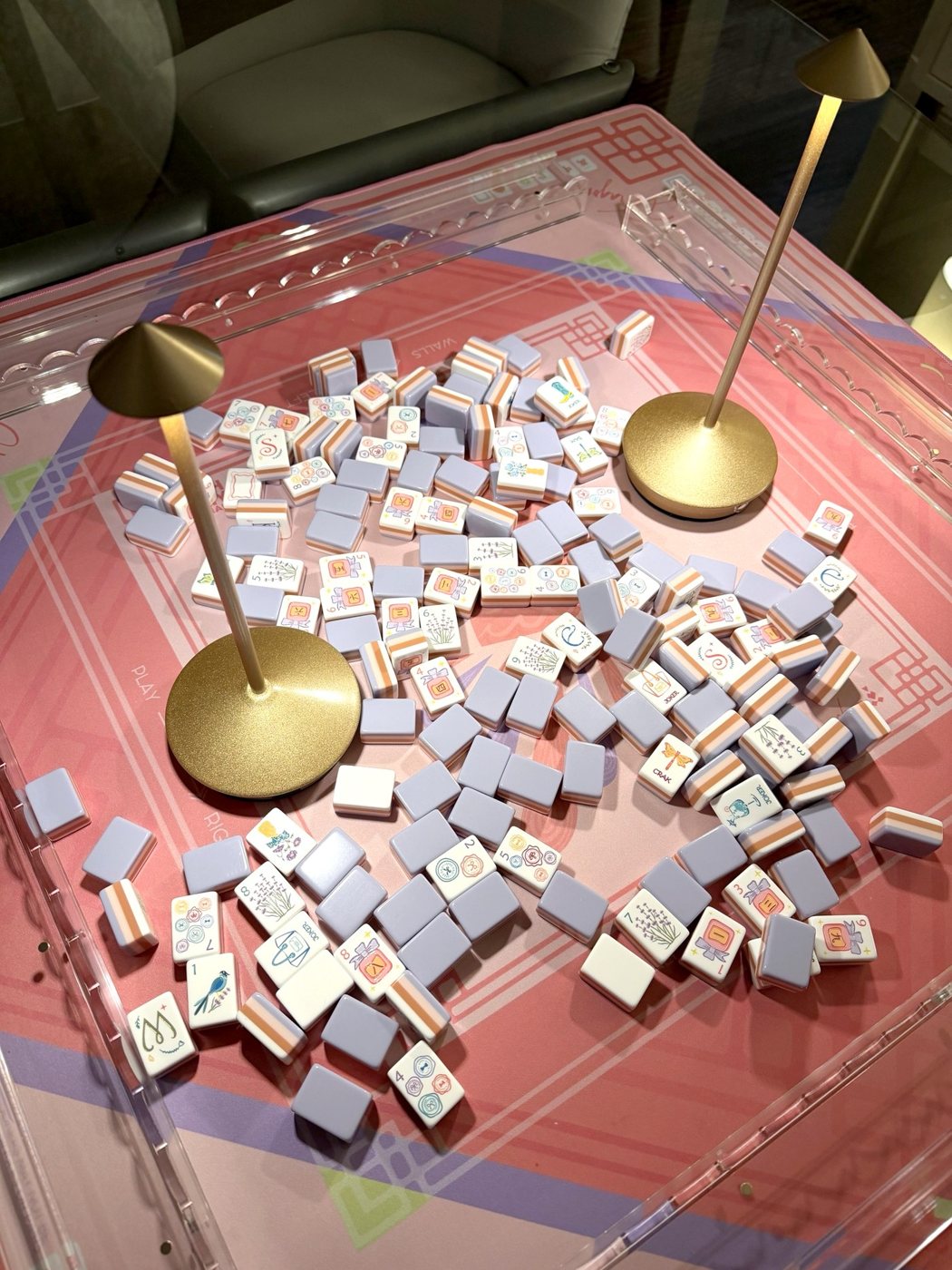 Two gold cordless table lamps glowing over purple and white mahjong tiles on a pink mat inside clear acrylic racks