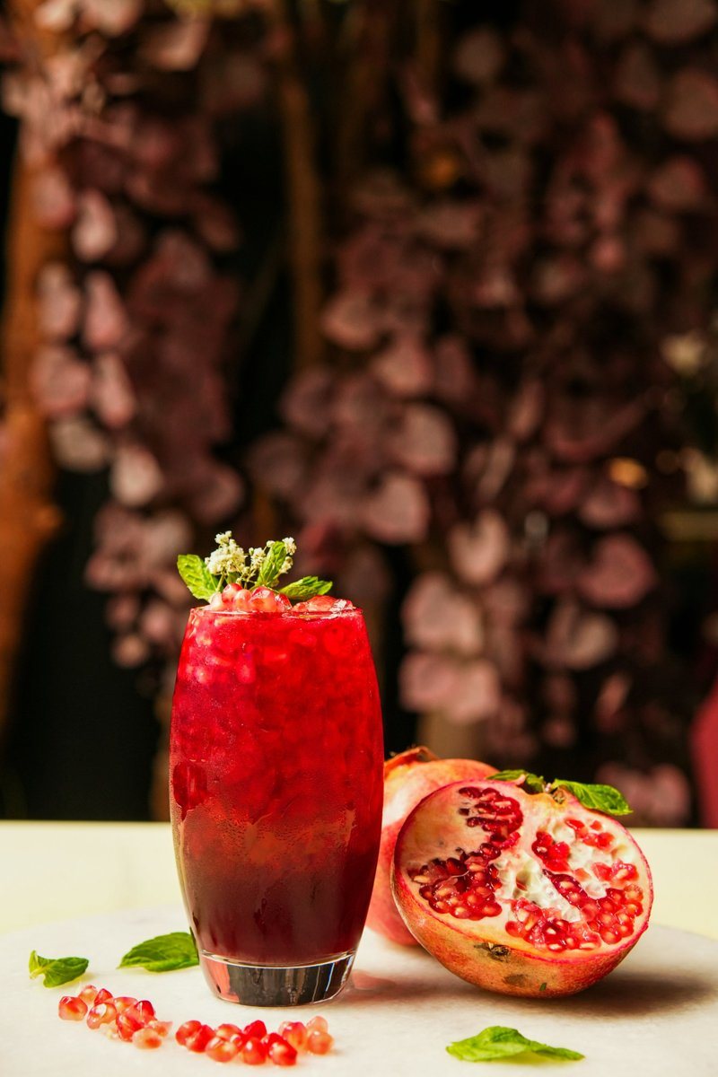 A tall ruby red pomegranate cocktail topped with pomegranate seeds and mint, beside a halved pomegranate